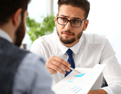 Ein Berater im weißen Hemd und mit Krawatte erklärt einem Kunden ein Dokument mit Diagrammen und Finanzdaten. Die Szene symbolisiert ein Beratungsgespräch über Kreditvoraussetzungen wie Bonität, Einkommen oder Sicherheiten.