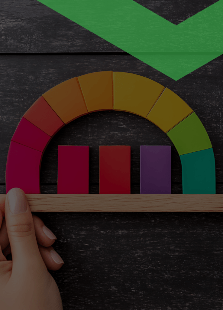 Eine Hand hält ein buntes Halbkreis-Diagramm aus Holz, das von Rot über Gelb zu Grün verläuft. In der Mitte stehen vier unterschiedlich hohe Balken. Die Szene symbolisiert die Überprüfung der Bonität, also der Kreditwürdigkeit einer Person oder eines Unternehmens.