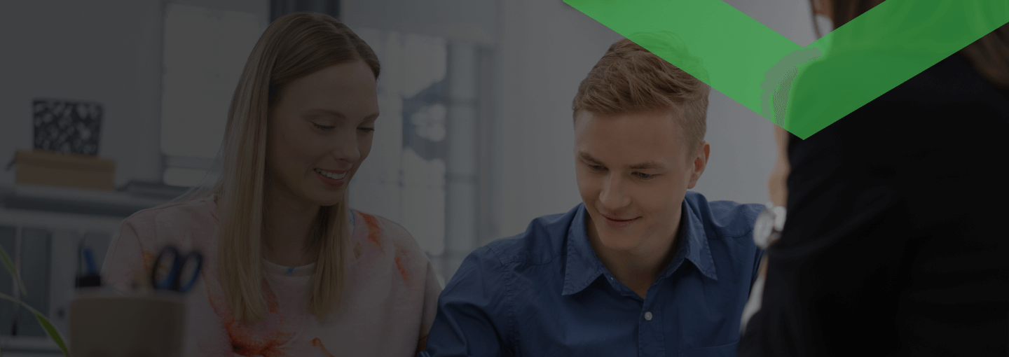 Ein junges Paar sitzt gemeinsam an einem Tisch und unterschreibt ein Dokument in Anwesenheit einer dritten Person. Die Szene symbolisiert den Abschluss eines Kredits mit Bürgschaft, bei dem ein Bürge zur zusätzlichen Absicherung hinzugezogen wird.