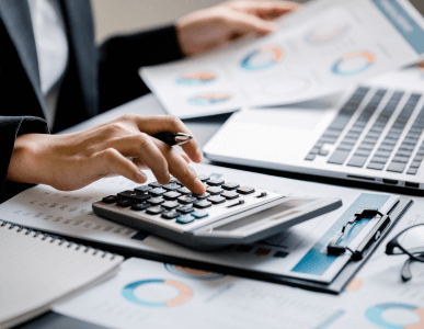 Eine Person arbeitet mit einem Taschenrechner und analysiert Finanzdiagramme, die auf dem Tisch und am Laptop sichtbar sind. Die Szene symbolisiert die Prüfung der Zahlungsfähigkeit, etwa im Rahmen einer Kreditwürdigkeitsbewertung oder Haushaltsrechnung.