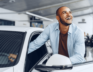 Ein junger Mann steht lächelnd neben einem weißen Auto und hält die Fahrertür offen. Die Szene symbolisiert die Übergabe oder Nutzung eines Fahrzeugs, etwa im Zusammenhang mit einem Eigentumsvorbehalt, bei dem das Auto erst mit vollständiger Zahlung in das Eigentum des Käufers übergeht.