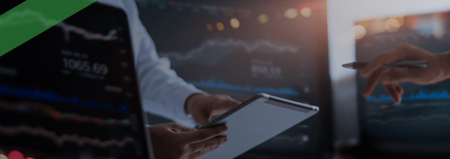Eine Person hält ein Tablet in der Hand und beobachtet Finanz- oder Börsendaten auf einem großen Bildschirm im Hintergrund. Im Vordergrund ist ein Laptop mit ähnlichen Kursgrafiken zu sehen. Die Szene symbolisiert Aktivitäten auf dem Kreditmarkt oder die Analyse von Finanz- und Kapitalmarktdaten.