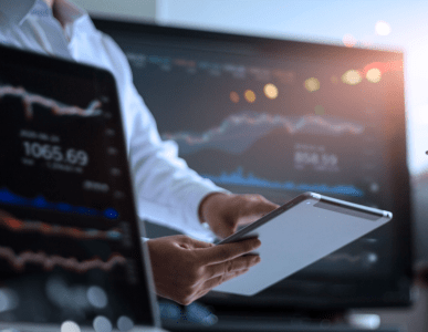 Eine Person hält ein Tablet in der Hand und beobachtet Finanz- oder Börsendaten auf einem großen Bildschirm im Hintergrund. Im Vordergrund ist ein Laptop mit ähnlichen Kursgrafiken zu sehen. Die Szene symbolisiert Aktivitäten auf dem Kreditmarkt oder die Analyse von Finanz- und Kapitalmarktdaten.