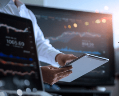 Eine Person hält ein Tablet in der Hand und beobachtet Finanz- oder Börsendaten auf einem großen Bildschirm im Hintergrund. Im Vordergrund ist ein Laptop mit ähnlichen Kursgrafiken zu sehen. Die Szene symbolisiert Aktivitäten auf dem Kreditmarkt oder die Analyse von Finanz- und Kapitalmarktdaten.