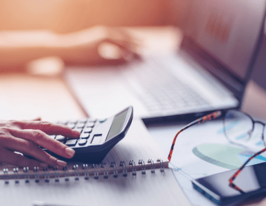 Eine Person tippt auf einem Taschenrechner, während im Hintergrund ein Laptop geöffnet ist. Auf dem Tisch liegen ein Notizbuch, ein Smartphone, eine Brille und Diagramme. Die Szene symbolisiert die Berechnung und Planung variabler Kreditkonditionen, etwa bei einem Cap-Darlehen mit Zinsobergrenze.