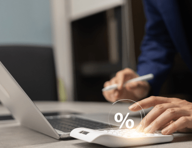 Eine Person tippt auf einem weißen Taschenrechner mit eingeblendetem Prozentzeichen, während sie mit der anderen Hand Notizen am Laptop macht. Die Szene symbolisiert die Berechnung des Nominalzinses, also des reinen Zinssatzes ohne Berücksichtigung weiterer Kosten.