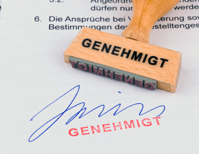 Ein Holzstempel mit der Aufschrift „GENEHMIGT“ liegt auf einem unterschriebenen Dokument, auf dem ebenfalls „GENEHMIGT“ in roter Farbe gestempelt wurde. Die Szene symbolisiert die offizielle Genehmigung oder Freigabe eines Antrags oder Vertrags.