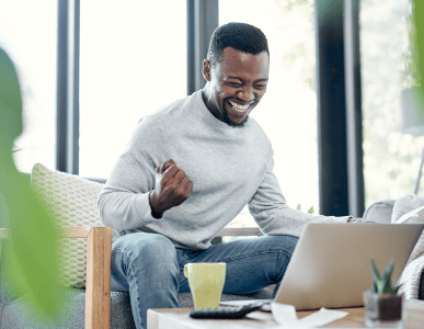 Ein Mann sitzt freudestrahlend vor seinem Laptop in einem gemütlichen Wohnzimmer, ballt die Faust vor Freude. Auf dem Tisch liegen Unterlagen, ein Taschenrechner und eine Tasse. Die Szene symbolisiert Erleichterung und Befreiung – etwa im Zusammenhang mit einer erfolgreichen Restschuldbefreiung.