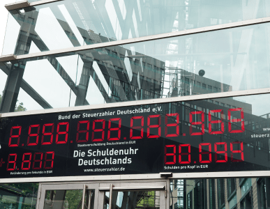 Die Schuldenuhr des Bundes der Steuerzahler Deutschlands ist an der Glasfassade eines Gebäudes angebracht und zeigt in roten Ziffern die aktuelle Staatsverschuldung sowie die Schulden pro Kopf an. Die Szene symbolisiert das Ausmaß der öffentlichen Verschuldung in Deutschland.