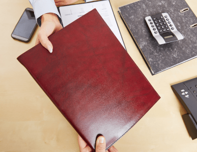 Zwei Personen reichen sich über einem Schreibtisch eine rote Dokumentenmappe. Auf dem Tisch liegen ein Taschenrechner, ein Ordner, ein Telefon und Unterlagen. Die Szene symbolisiert die Übergabe von Forderungsunterlagen im Rahmen einer Zession, also einer Abtretung von Ansprüchen.