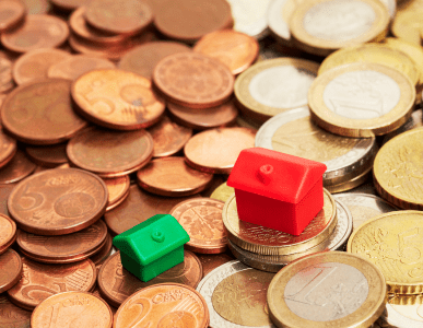 Zwei unterschiedlich große Spielzeughäuser stehen auf einem Haufen Euro-Münzen. Die Szene symbolisiert die Zinsspanne, also die Differenz zwischen Soll- und Habenzinsen bei der Kreditvergabe durch Banken.