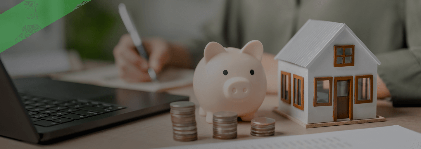 Ein Sparschwein, gestapelte Münzen und ein Modellhaus stehen auf einem Schreibtisch mit Finanzunterlagen. Im Hintergrund schreibt eine Person etwas auf. Die Szene symbolisiert eine Anzahlung im Rahmen eines Immobilienkaufs oder einer Finanzierung.