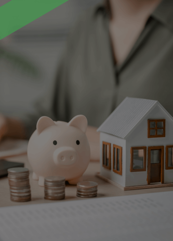 Ein Sparschwein, gestapelte Münzen und ein Modellhaus stehen auf einem Schreibtisch mit Finanzunterlagen. Im Hintergrund schreibt eine Person etwas auf. Die Szene symbolisiert eine Anzahlung im Rahmen eines Immobilienkaufs oder einer Finanzierung.