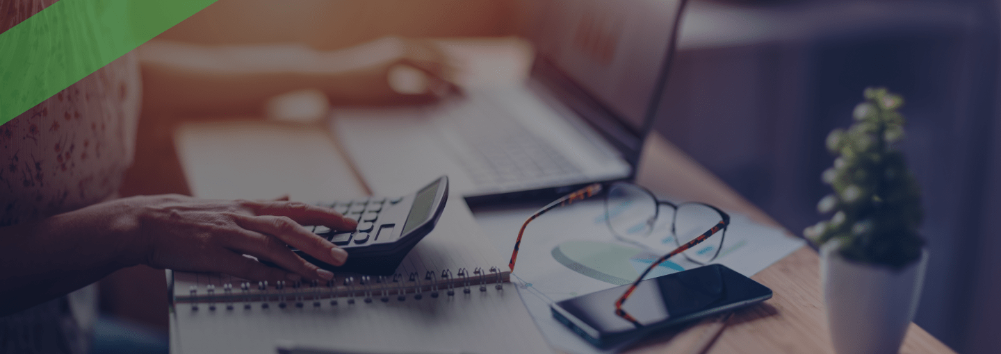 Eine Person tippt auf einem Taschenrechner, während im Hintergrund ein Laptop geöffnet ist. Auf dem Tisch liegen ein Notizbuch, ein Smartphone, eine Brille und Diagramme. Die Szene symbolisiert die Berechnung und Planung variabler Kreditkonditionen, etwa bei einem Cap-Darlehen mit Zinsobergrenze.