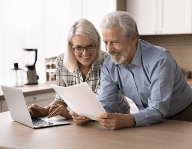 Ein älteres Paar steht in einer hellen Küche, schaut gemeinsam lächelnd auf ein Dokument und den Laptop. Die Szene symbolisiert die gemeinsame Prüfung oder Planung eines Darlehensbetrags, etwa im Rahmen einer Finanzierung oder Umschuldung.