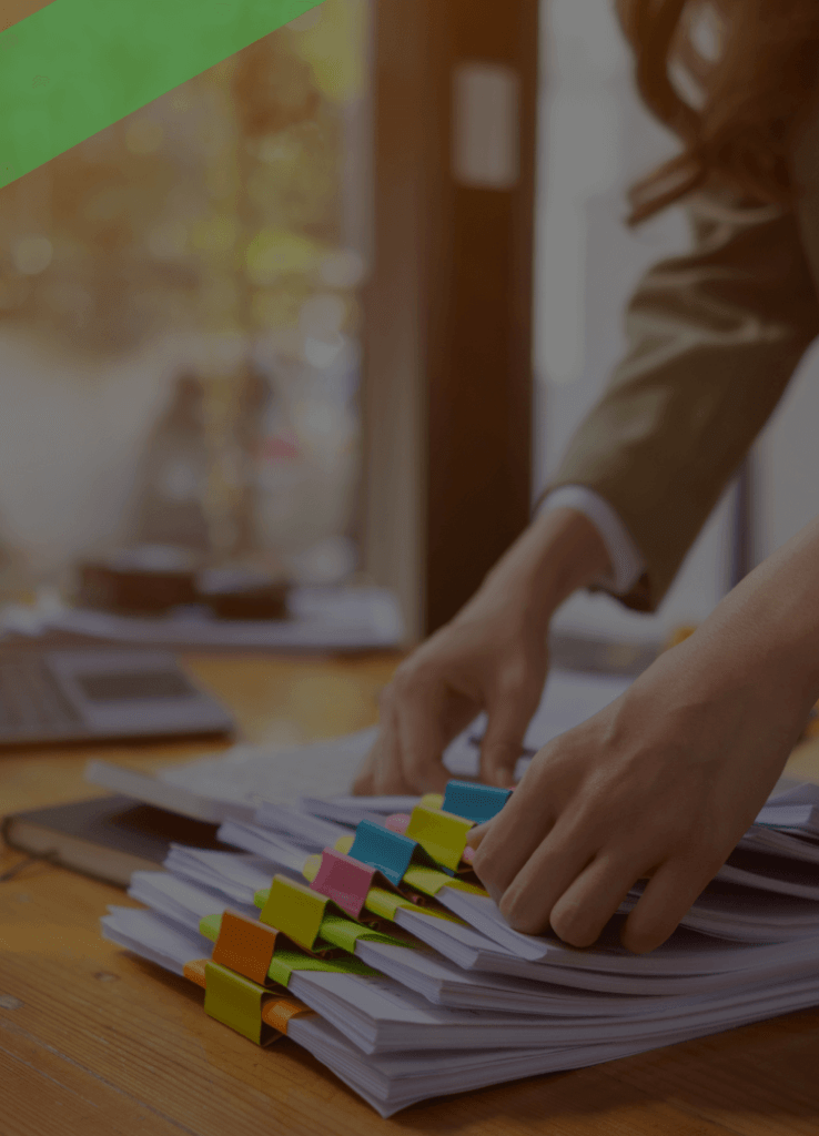 Eine Frau sortiert mehrere Dokumentenstapel mit farbigen Markierungen auf einem Schreibtisch. Im Hintergrund steht ein Laptop. Die Szene symbolisiert das Management von Debitoren, also offenen Forderungen gegenüber Kunden oder Kreditnehmern.