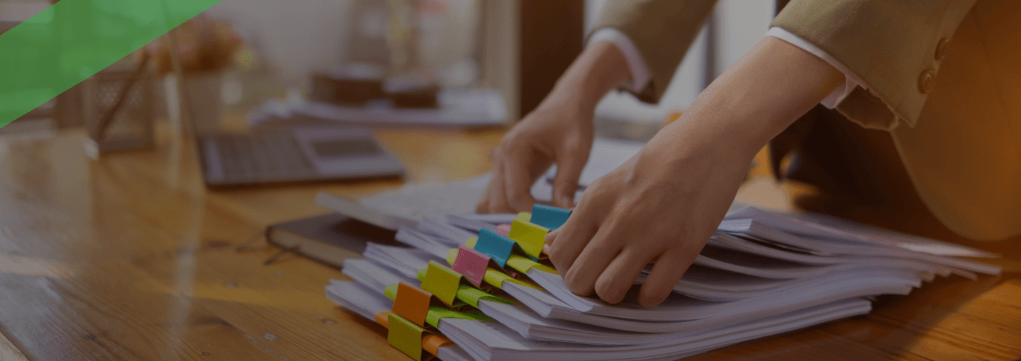 Eine Frau sortiert mehrere Dokumentenstapel mit farbigen Markierungen auf einem Schreibtisch. Im Hintergrund steht ein Laptop. Die Szene symbolisiert das Management von Debitoren, also offenen Forderungen gegenüber Kunden oder Kreditnehmern.
