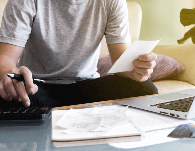 Ein Mann sitzt auf einem Sofa und überprüft mit einem Taschenrechner und Belegen seine Unterlagen. Auf dem Tisch liegen ein Laptop, Quittungen, ein Notizbuch und eine Kreditkarte. Die Szene symbolisiert die Zusammenstellung eines Einkommensnachweises zur Vorlage bei einer Finanzierung oder einem Kreditantrag.