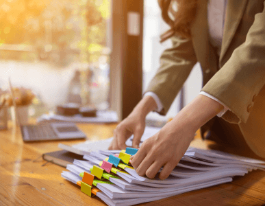 Eine Frau sortiert mehrere Dokumentenstapel mit farbigen Markierungen auf einem Schreibtisch. Im Hintergrund steht ein Laptop. Die Szene symbolisiert das Management von Debitoren, also offenen Forderungen gegenüber Kunden oder Kreditnehmern.
