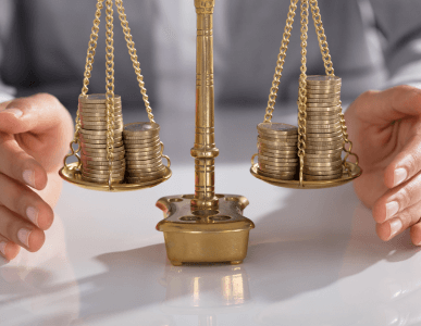 Eine klassische Waage mit gestapelten Münzen auf beiden Seiten steht auf einem Tisch, im Hintergrund hält eine Person die Waage vorsichtig. Die Szene symbolisiert finanzielle Abwägungen, etwa im Zusammenhang mit einem Disagio – dem Abschlag vom Nennwert eines Kredits bei Auszahlung.