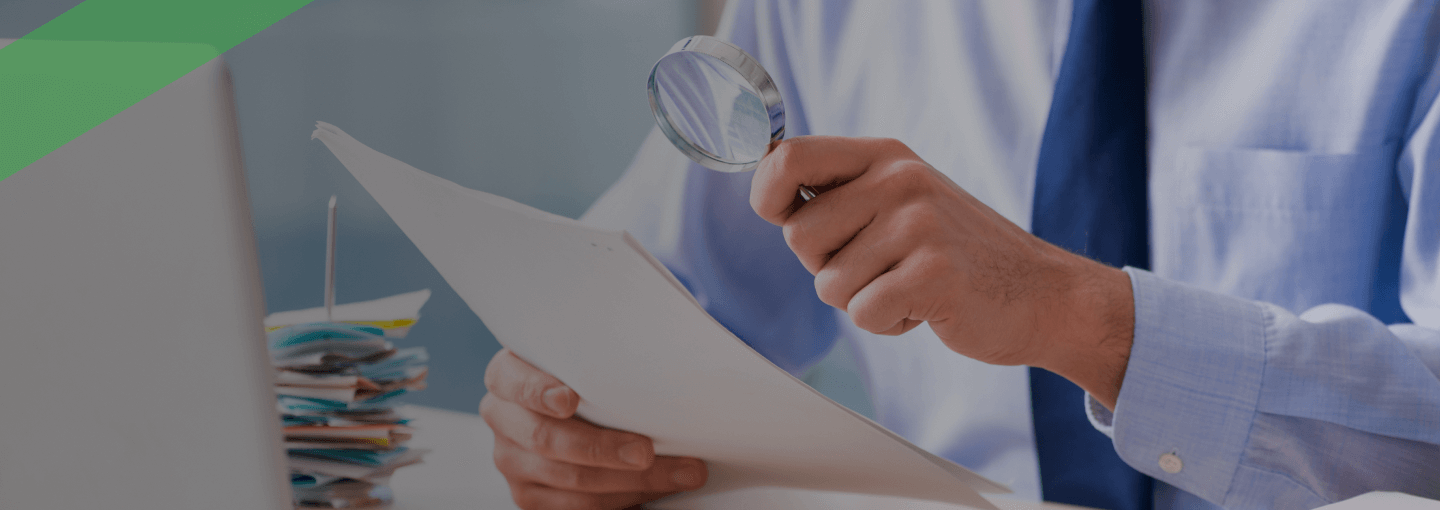 Ein Mann in Businesskleidung sitzt am Schreibtisch und betrachtet aufmerksam ein Dokument mit einer Lupe. Vor ihm liegen ein Laptop, ein Taschenrechner und mehrere Aktenstapel. Die Szene symbolisiert die sorgfältige Prüfung von Unterlagen im Zusammenhang mit der Aufdeckung oder Vermeidung von Kreditbetrug.