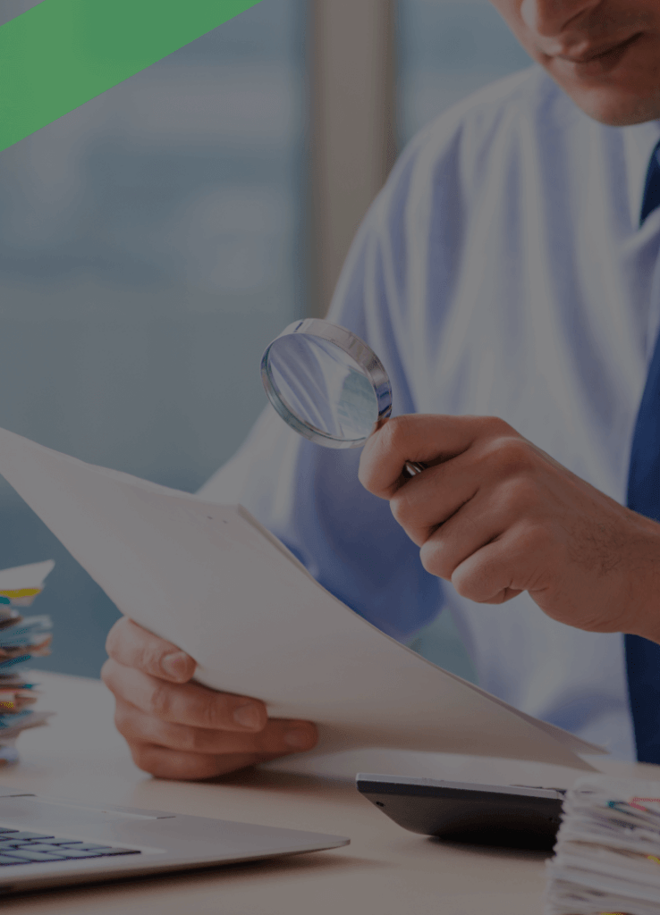 Ein Mann in Businesskleidung sitzt am Schreibtisch und betrachtet aufmerksam ein Dokument mit einer Lupe. Vor ihm liegen ein Laptop, ein Taschenrechner und mehrere Aktenstapel. Die Szene symbolisiert die sorgfältige Prüfung von Unterlagen im Zusammenhang mit der Aufdeckung oder Vermeidung von Kreditbetrug.