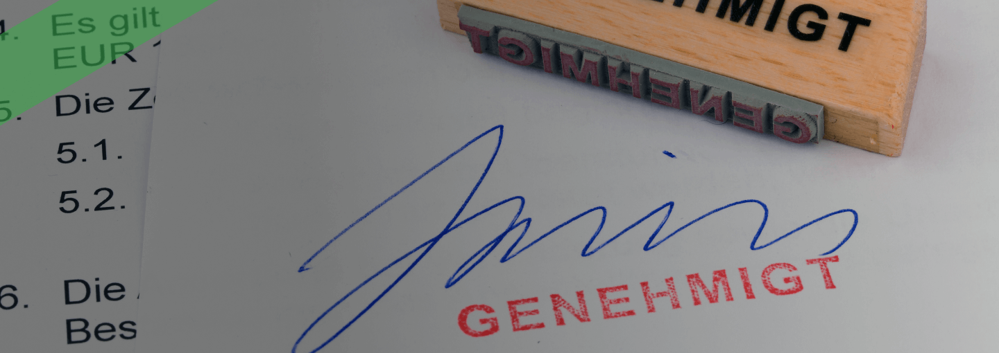Ein Holzstempel mit der Aufschrift „GENEHMIGT“ liegt auf einem unterschriebenen Dokument, auf dem ebenfalls „GENEHMIGT“ in roter Farbe gestempelt wurde. Die Szene symbolisiert die offizielle Genehmigung oder Freigabe eines Antrags oder Vertrags.