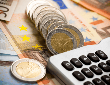 Mehrere gestapelte Euromünzen liegen auf Euro-Banknoten neben einem Taschenrechner. Die Szene symbolisiert finanzielle Kalkulationen im Zusammenhang mit kurzfristigen Krediten oder Liquiditätsengpässen.