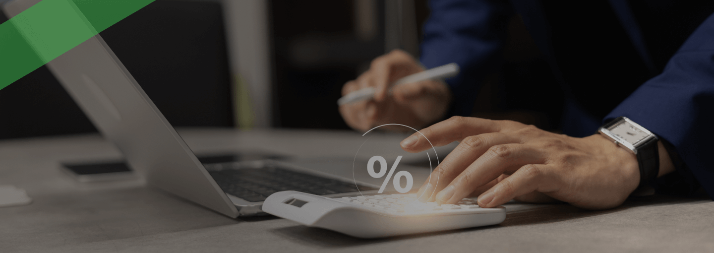 Eine Person tippt auf einem weißen Taschenrechner mit eingeblendetem Prozentzeichen, während sie mit der anderen Hand Notizen am Laptop macht. Die Szene symbolisiert die Berechnung des Nominalzinses, also des reinen Zinssatzes ohne Berücksichtigung weiterer Kosten.