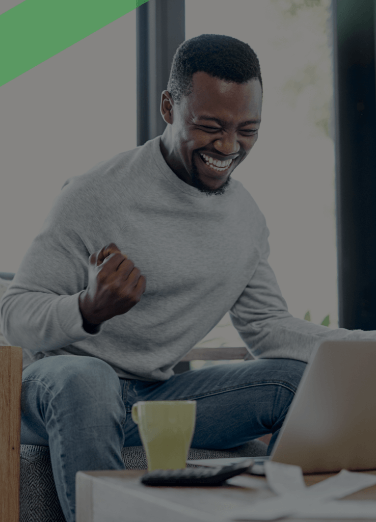 Ein Mann sitzt freudestrahlend vor seinem Laptop in einem gemütlichen Wohnzimmer, ballt die Faust vor Freude. Auf dem Tisch liegen Unterlagen, ein Taschenrechner und eine Tasse. Die Szene symbolisiert Erleichterung und Befreiung – etwa im Zusammenhang mit einer erfolgreichen Restschuldbefreiung.