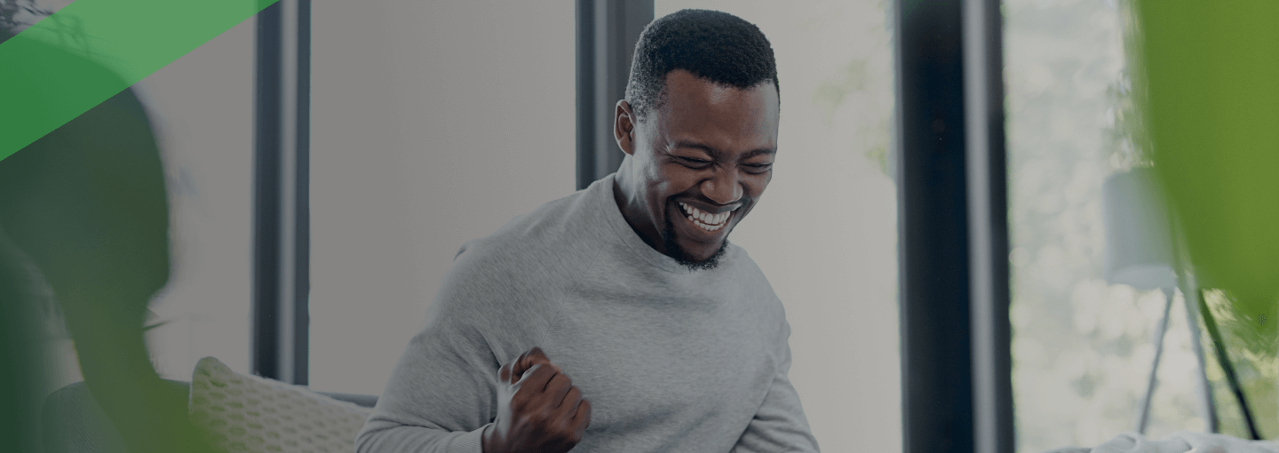 Ein Mann sitzt freudestrahlend vor seinem Laptop in einem gemütlichen Wohnzimmer, ballt die Faust vor Freude. Auf dem Tisch liegen Unterlagen, ein Taschenrechner und eine Tasse. Die Szene symbolisiert Erleichterung und Befreiung – etwa im Zusammenhang mit einer erfolgreichen Restschuldbefreiung.