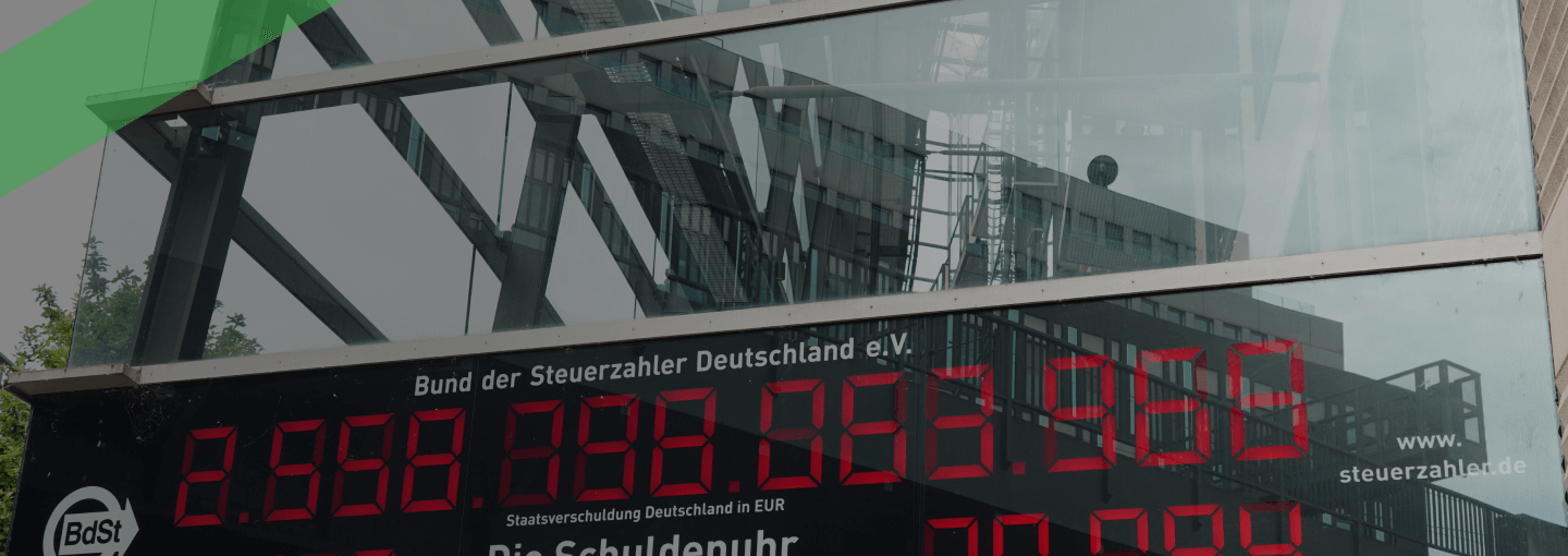 Die Schuldenuhr des Bundes der Steuerzahler Deutschlands ist an der Glasfassade eines Gebäudes angebracht und zeigt in roten Ziffern die aktuelle Staatsverschuldung sowie die Schulden pro Kopf an. Die Szene symbolisiert das Ausmaß der öffentlichen Verschuldung in Deutschland.