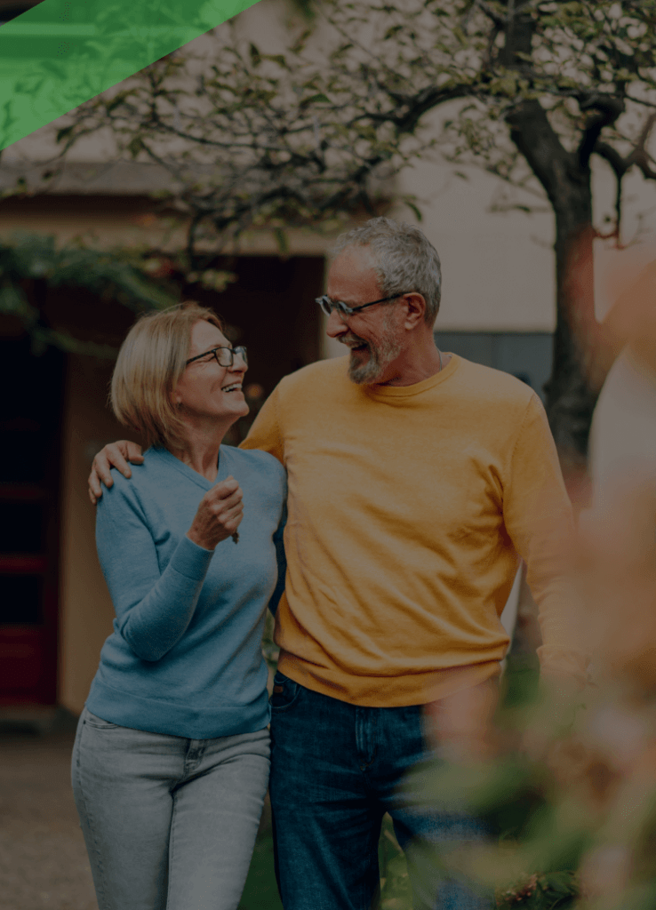 Ein älteres Paar steht lachend Arm in Arm im Vorgarten vor einem Wohnhaus. Die Szene symbolisiert finanzielle Unabhängigkeit im Alter und steht sinnbildlich für eine Umkehrhypothek, bei der Immobilieneigentum in regelmäßige Zahlungen umgewandelt wird.
