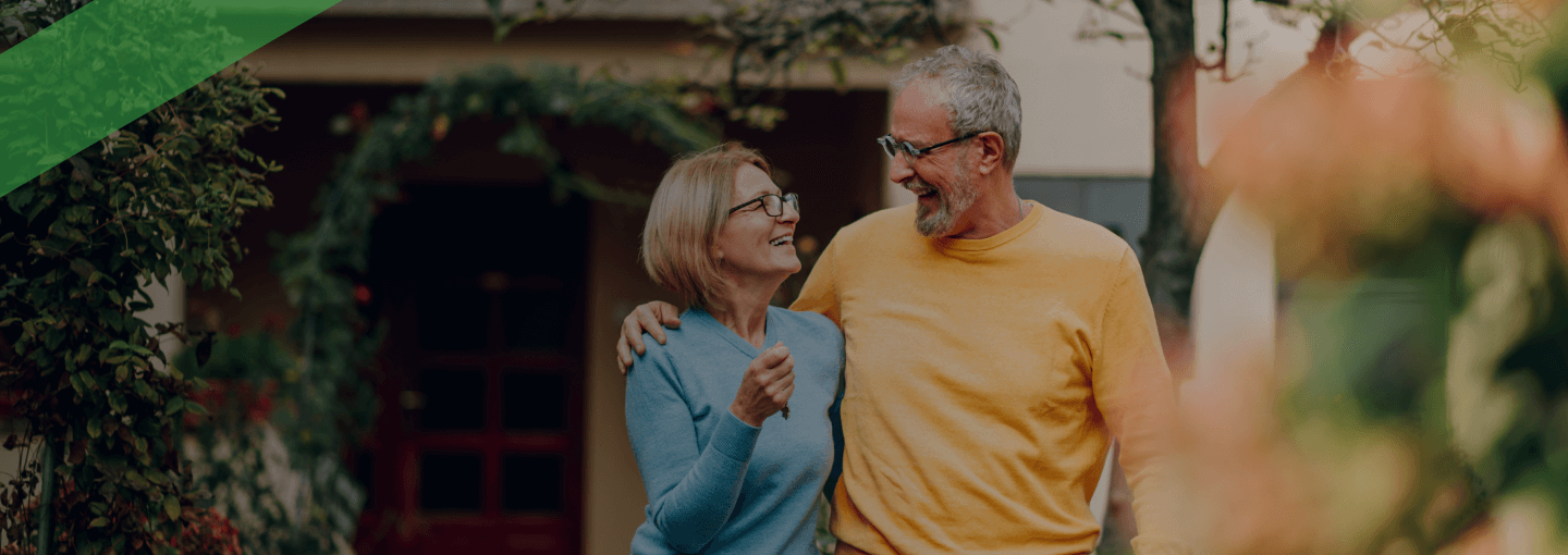 Ein älteres Paar steht lachend Arm in Arm im Vorgarten vor einem Wohnhaus. Die Szene symbolisiert finanzielle Unabhängigkeit im Alter und steht sinnbildlich für eine Umkehrhypothek, bei der Immobilieneigentum in regelmäßige Zahlungen umgewandelt wird.