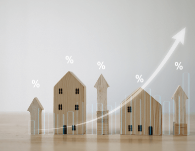 Mehrere unterschiedlich große Holzhaus-Modelle stehen nebeneinander. Darüber sind Prozentzeichen sowie ein ansteigender Pfeil mit transparenten Balkendiagrammen eingeblendet. Die Darstellung symbolisiert steigende Zinssätze in Abhängigkeit von Faktoren wie Bonität oder Immobilienwert.