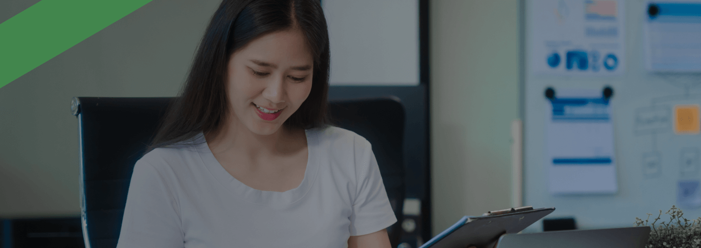 Eine junge Frau sitzt an einem Schreibtisch, hält einen Stift und ein Klemmbrett in der Hand und lächelt, während sie Unterlagen durchgeht. Auf dem Tisch liegen ein Laptop und Aktenordner. Die Szene symbolisiert die Bearbeitung oder Bewertung von Finanz- oder Bonitätsunterlagen, etwa im Zusammenhang mit einem Bonitätsindex.