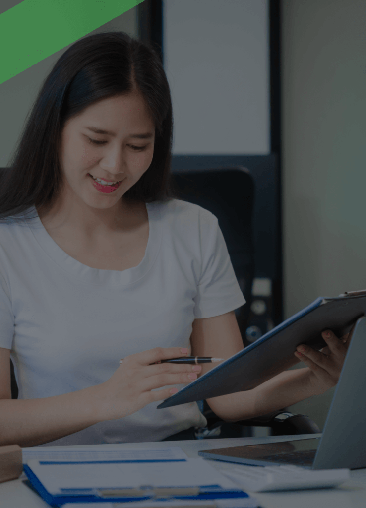 Eine junge Frau sitzt an einem Schreibtisch, hält einen Stift und ein Klemmbrett in der Hand und lächelt, während sie Unterlagen durchgeht. Auf dem Tisch liegen ein Laptop und Aktenordner. Die Szene symbolisiert die Bearbeitung oder Bewertung von Finanz- oder Bonitätsunterlagen, etwa im Zusammenhang mit einem Bonitätsindex.