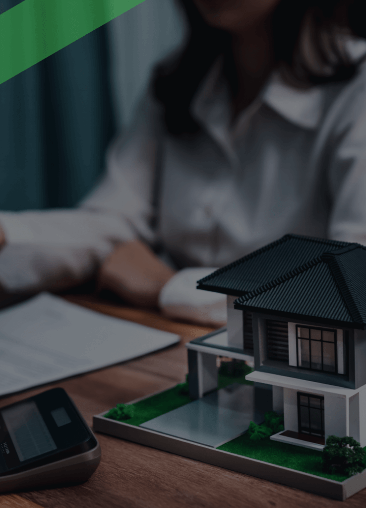 Ein Berater übergibt einer Kundin einen Schlüssel über einem Schreibtisch, auf dem ein Vertrag, ein Taschenrechner und ein Modellhaus liegen. Die Szene symbolisiert den Abschluss einer Immobilienfinanzierung, etwa im Zusammenhang mit einer endfälligen Darlehensrückzahlung.