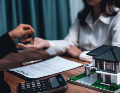 Ein Berater übergibt einer Kundin einen Schlüssel über einem Schreibtisch, auf dem ein Vertrag, ein Taschenrechner und ein Modellhaus liegen. Die Szene symbolisiert den Abschluss einer Immobilienfinanzierung, etwa im Zusammenhang mit einer endfälligen Darlehensrückzahlung.