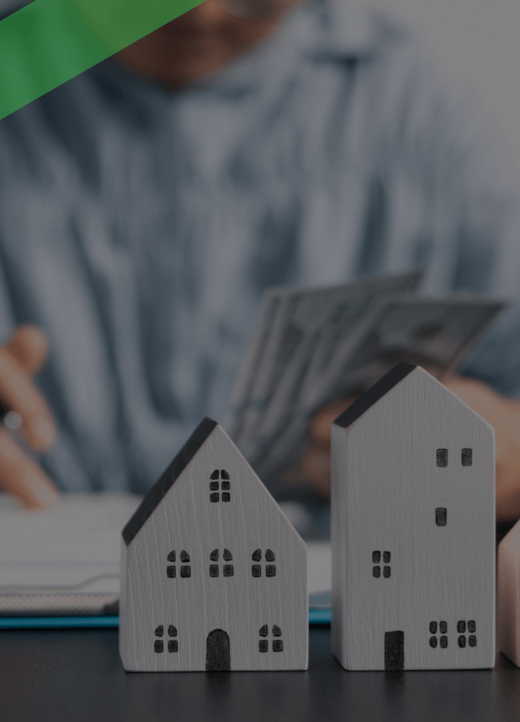 Im Vordergrund stehen vier kleine Modellhäuser aus Holz aufgereiht nebeneinander. Im Hintergrund zählt eine Person Geldscheine. Die Szene symbolisiert finanzielle Absicherung im Immobilienbereich, etwa durch einen Garantiekredit oder eine Bürgschaft.