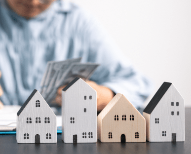 Im Vordergrund stehen vier kleine Modellhäuser aus Holz aufgereiht nebeneinander. Im Hintergrund zählt eine Person Geldscheine. Die Szene symbolisiert finanzielle Absicherung im Immobilienbereich, etwa durch einen Garantiekredit oder eine Bürgschaft.