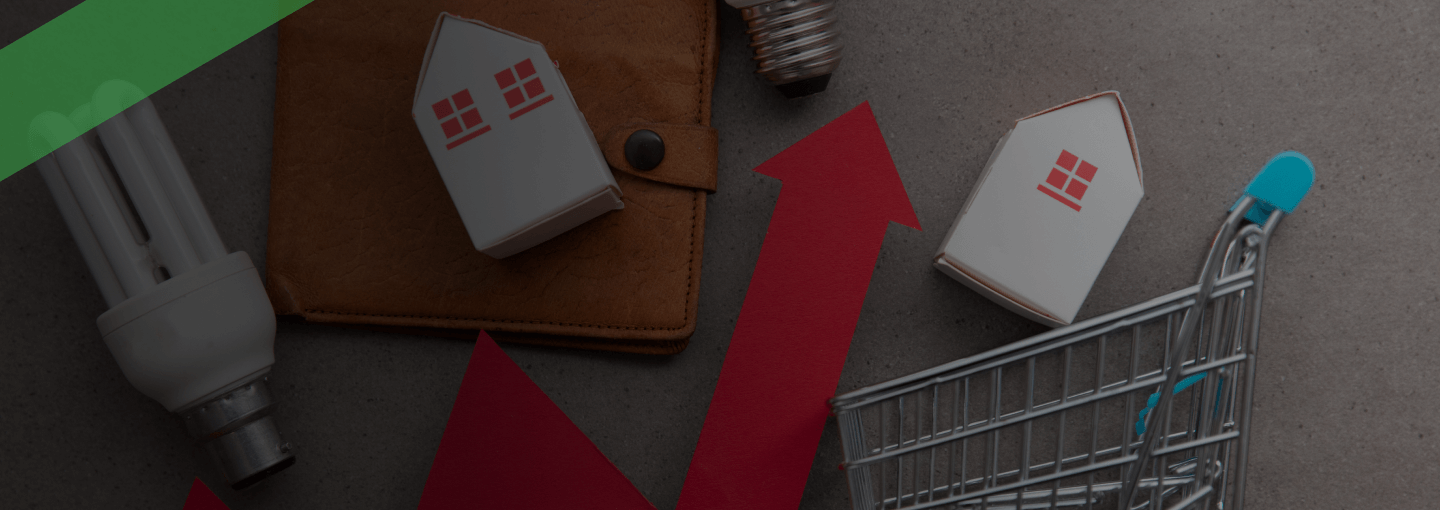 Eine rote Zickzacklinie mit Pfeil nach oben verläuft zwischen Symbolen für Konsum und Wohnen: Miniaturhäuser, ein Einkaufswagen, Glühbirnen und eine Geldbörse. Die Szene symbolisiert Inflation und den Anstieg der Lebenshaltungskosten.