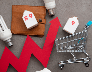 Eine rote Zickzacklinie mit Pfeil nach oben verläuft zwischen Symbolen für Konsum und Wohnen: Miniaturhäuser, ein Einkaufswagen, Glühbirnen und eine Geldbörse. Die Szene symbolisiert Inflation und den Anstieg der Lebenshaltungskosten.