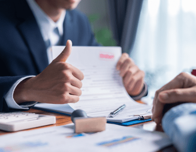Ein Berater in Anzug hält ein Vertragsdokument und zeigt mit dem Daumen nach oben. Auf dem Tisch liegen Unterlagen, ein Stempel, ein Taschenrechner und ein Klemmbrett. Die Szene symbolisiert die Genehmigung oder den Abschluss eines Investitionskredits zur Finanzierung geschäftlicher Vorhaben.