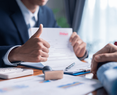 Ein Berater in Anzug hält ein Vertragsdokument und zeigt mit dem Daumen nach oben. Auf dem Tisch liegen Unterlagen, ein Stempel, ein Taschenrechner und ein Klemmbrett. Die Szene symbolisiert die Genehmigung oder den Abschluss eines Investitionskredits zur Finanzierung geschäftlicher Vorhaben.