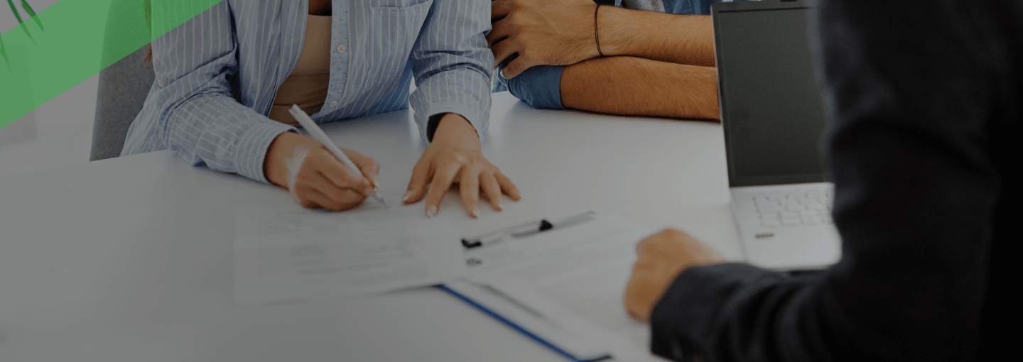 Ein junges Paar sitzt lächelnd an einem Tisch und unterzeichnet gemeinsam einen Vertrag, während ein Berater ihnen gegenüber sitzt. Auf dem Tisch liegen weitere Unterlagen und ein Laptop. Die Szene symbolisiert den Abschluss eines Kreditvertrags oder einer Finanzierung.