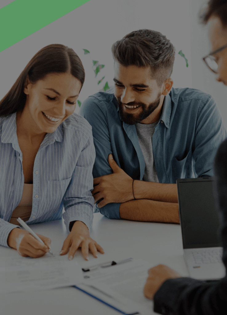 Ein junges Paar sitzt lächelnd an einem Tisch und unterzeichnet gemeinsam einen Vertrag, während ein Berater ihnen gegenüber sitzt. Auf dem Tisch liegen weitere Unterlagen und ein Laptop. Die Szene symbolisiert den Abschluss eines Kreditvertrags oder einer Finanzierung.