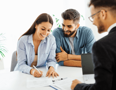 Ein junges Paar sitzt lächelnd an einem Tisch und unterzeichnet gemeinsam einen Vertrag, während ein Berater ihnen gegenüber sitzt. Auf dem Tisch liegen weitere Unterlagen und ein Laptop. Die Szene symbolisiert den Abschluss eines Kreditvertrags oder einer Finanzierung.