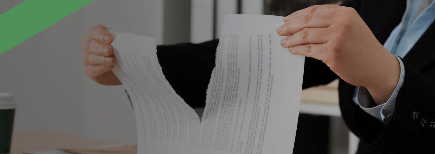 Eine Person zerreißt einen Vertrag oder ein Dokument in der Mitte über einem Schreibtisch. Auf dem Tisch befinden sich eine Klemmbrettmappe mit weiteren Unterlagen sowie Büromaterialien im Hintergrund. Die Szene deutet auf die Kündigung oder Auflösung eines Vertrages hin.