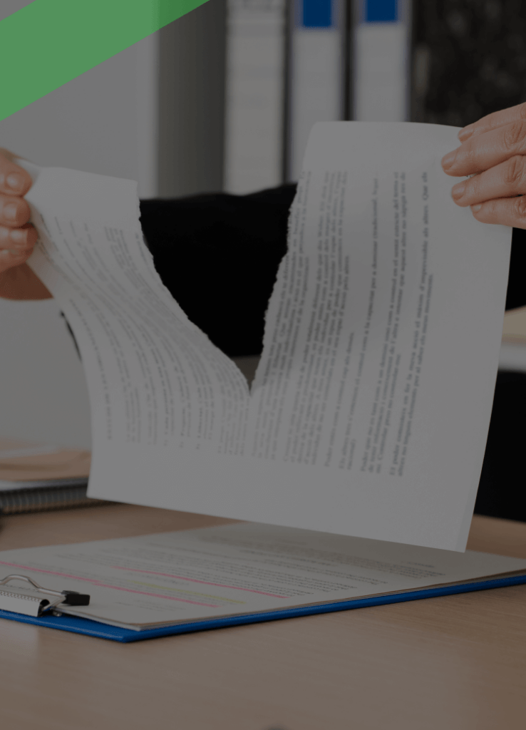 Eine Person zerreißt einen Vertrag oder ein Dokument in der Mitte über einem Schreibtisch. Auf dem Tisch befinden sich eine Klemmbrettmappe mit weiteren Unterlagen sowie Büromaterialien im Hintergrund. Die Szene deutet auf die Kündigung oder Auflösung eines Vertrages hin.
